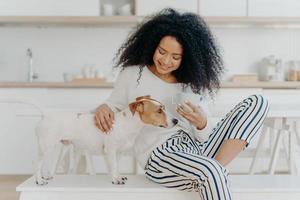 Lovely African American woman has coffee break, petting pedigree dog, sits on comfortable white bench against kitchen background, smiles with happiness. People, care, love and animals concept photo