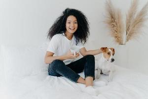 Happy relaxed lovely young woman with Afro hairstyle, petting dog, sit together on bed with white bedclothes, drink hot beverage, dressed in casual wear, expresses positive emotions. People and rest photo