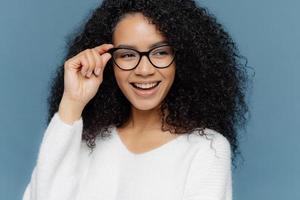 primer plano de una mujer alegre que usa anteojos, mantiene la mano en el marco, mira alegremente a un lado, nota algo asombroso en la distancia, vestida con un jersey blanco informal, modelos con fondo azul foto