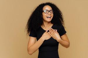 Thankful positive woman smiles happily, makes gratitude gesture, keeps hands on chest, smiles broadly, wears casual t shirt, optical glasses, closes eyes from pleasure, isolated on beige background photo
