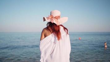 mooi vrouw in een wit overhemd en hoed wandelingen langs de strand naar de zee Aan een helder zonnig dag. zomer tijd, vakantie, rust uit. langzaam beweging.