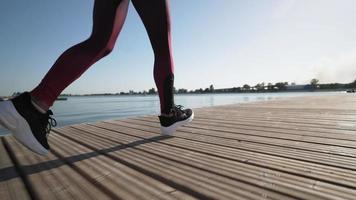 Ein junges athletisches Mädchen läuft in schwarzen Turnschuhen und Leggings auf einem Pantone am See entlang. Zeitlupe. Nahansicht. Beine. video