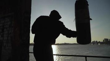 Silhouette of a boxer against the backdrop of a beautiful sunset and a river in an abandoned building, trains blows on a punching bag. Epic video. Motivation. Sports and martial arts. Slow motion. video