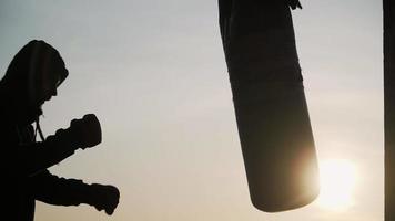 el boxeador recortado contra el telón de fondo de una hermosa puesta de sol golpea un saco de boxeo rápidamente hasta el fracaso. fatiga, ya no puede hacer ejercicio. motivación. deportes y artes marciales. video