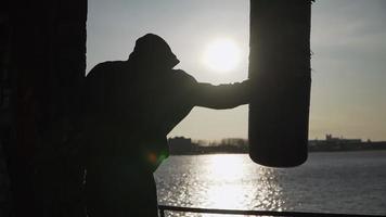 Silhouette of a boxer against the backdrop of a beautiful sunset and a river in an abandoned building, trains blows on a punching bag. Epic video. Motivation. Sports and martial arts. Slow motion. video