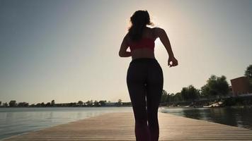 uma jovem atlética corre ao longo do pantone ao longo do lago em tênis pretos e leggings contra o pôr do sol. câmera lenta. plano geral. video