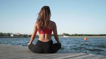 Young athletic Caucasian girl in a red tank top practices yoga on a lake background. Sits in a lotus position on a pantone. video