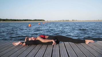 una joven atlética caucásica con una camiseta roja y polainas está haciendo estiramientos y yoga en el fondo del lago. flexibilidad. video