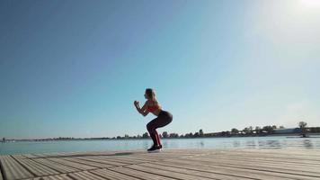 jeune fille caucasienne athlétique dans un débardeur rouge faisant des squats sur un fond de lac. il est engagé dans le fitness. video