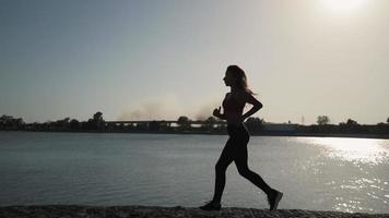 giovane atletico caucasico ragazza corre lungo il lago su un' tramonto sfondo. lontano lontano Fumo a partire dal il fuoco. lento movimento. complessivamente Piano. video