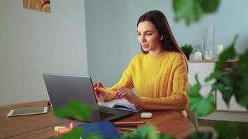 aluna e aprendendo usando o bate-papo da webcam do laptop faz anotações e trabalha em casa. jovem bonita de suéter amarelo aprende on-line com um professor por teleconferência. conceito de educação a distância. video