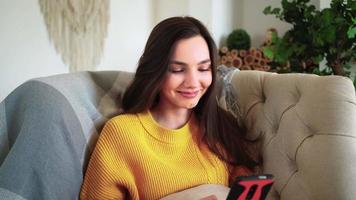 rilassato donna nel giallo maglione sorridente hold smartphone Guardando sociale media storie video sedersi su divano casa. contento ragazza Guarda cellulare ridendo godendo utilizzando mobile App avendo divertimento giocare e chat