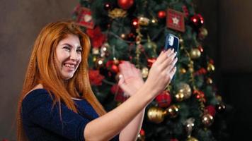 een mooi roodharig vrouw zit De volgende naar een Kerstmis boom versierd met flikkeren lichten en toepassingen haar smartphone naar communiceren van een afstand. luistert naar nieuw jaar hartelijk groeten, lacht en is gelukkig. video