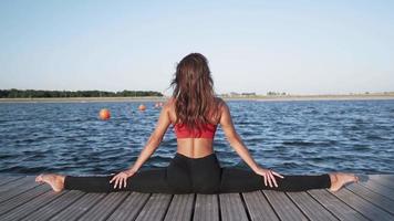 jeune fille caucasienne athlétique dans un débardeur rouge et des leggings est assise dans une scission sur fond de lac. engagé dans l'étirement. souplesse. video