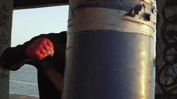 A boxer in a black hoodie on his head hood trains punches in red gloves on a punching bag. Abandoned building on the background. Motivation. Sports and martial arts. Close-up. Slow motion video