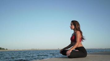 Junges athletisches kaukasisches Mädchen in einem roten Trägershirt praktiziert Yoga auf einem Seehintergrund. sitzt im Lotussitz auf einer Pantone. video