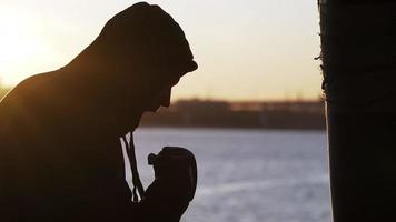 Silhouette of a boxer against the backdrop of a beautiful sunset and a river in an abandoned building, trains blows on a punching bag. Epic video. Motivation. Sports and martial arts. Slow motion. video