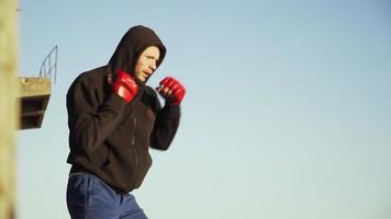 un tipo de apariencia caucásica, un boxeador con guantes rojos en una sudadera negra con capucha, hace un ejercicio de lucha en la sombra contra el fondo de un edificio abandonado. bajo tierra. camara lenta. video