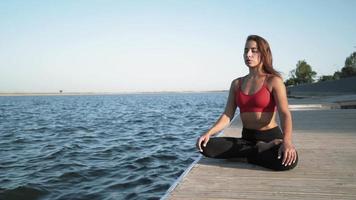 Young athletic Caucasian girl in a red tank top practices yoga on a lake background. Sits in a lotus position on a pantone. video