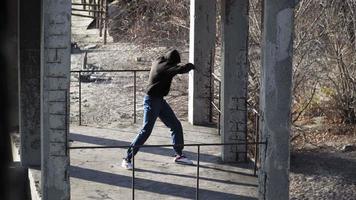 A guy Caucasian appearance A boxer black sweater with hood doing shadow boxing exercise against backdrop an abandoned building. Underground. Overall plan. Motivational video. video