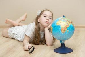 una niña pensativa yace en el suelo junto a un globo, sostiene una lupa en una mano, pone la otra debajo de su cabeza y discute. el concepto de niño inteligente, educación, planes para el futuro foto