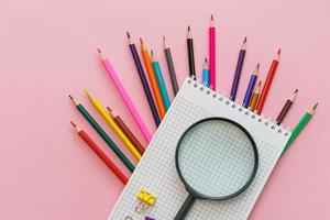 Magnifying glass, pencils and a blank white notepad on a spring on a pink photo