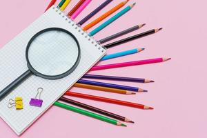 Magnifying glass, pencils and a blank white notepad on a spring on a pink photo