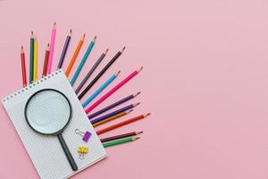 Magnifying glass, pencils and a blank white notepad on a spring on a pink photo
