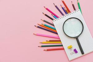 Magnifying glass, pencils and a blank white notepad on a spring on a pink photo