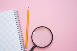 Magnifying glass, pencils and a blank white notepad on a spring on a pink photo