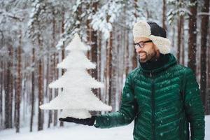 Smiling middle aged male looks with happy expression at artificial white small fir tree, going to celebrate New Years holidays, spends morning on fresh air in forest during snowy weather photo