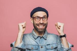 Joyful man with thick beard and mustache clenches fists and looks joyfully at camera, celebrates his victory, has happy expression, isolated over pink background. People, emotions and success photo