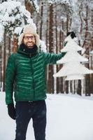 Portrait of cheerful bearded handsome male wears warm hat, green anorak and gloves holds little white artificial fir tree, stands in winter forest covered with snow. Beautiful landscapes photo