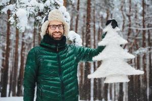 Pleasant looking bearded male dressed in warm clothes, holds artificial fir tree, stands against beautiful trees covered with snow, has cheerful positive expression, breathes fresh winter air photo