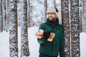 Happy bearded man in green jacket, stands near tree, holds firewood, looks thoughtfully aside, has cheerful expression, stands against snowy forest background. Fashionable pensive male outdoor photo