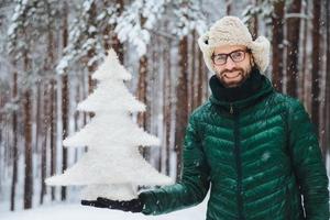 Smiling positive male wears glasses, hat and anorak, holds artificial fir tree, stands against trees covered with snow, looks directly into camera with happy expression, spends time on frosty weather photo