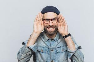 Portrait of happy hipster guy with thick beard and mustache wears stylish hat and glasses, keeps hands near face, tries to see something in darkness, isolated over grey background, has fun indoor photo