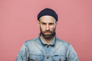 foto de un hombre barbudo de buen aspecto que parpadea y mira seriamente a la cámara, lleva una chaqueta vaquera y un sombrero negro, aislado sobre un fondo rosa. hombre elegante con poses de rastrojo en el estudio solo.