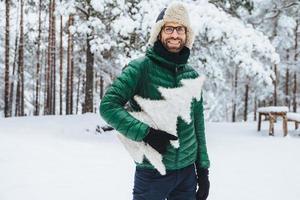 Pleasant looking bearded male wears warm clothes, holds white fir tree, stands against winter forest background, walks with friends during frosty weather, being in good mood. People and nature photo