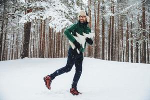 apuesto joven sonriente usa gorro y anorak, pasa tiempo libre al aire libre en el bosque durante el invierno, se prepara para las vacaciones, sostiene un abeto. un tipo barbudo con anteojos admira los paisajes invernales foto