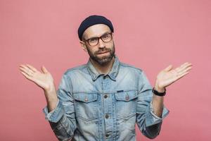 Portrait of hesitant bearded male with doubtful expression, shrugs shoulders, wears denim jacket, glasses and hat, isolated over pink studio background. Confused middle aged man poses indoor photo