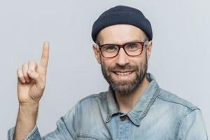 Positive handsome bearded male raises fore fimger, looks with cheerful expression directly intto camera, wears fashionable hat, shirt, raises fore finger as remebers something, models indoor photo