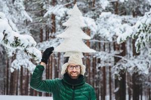 Funny bearded male with beard, dressed in warm winter clothes, holds artificial fir tree on head, poses against trees covered with snow, being in good mood. Optimistic man spends time outdoor photo