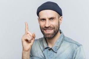 atractivo hombre de mediana edad levanta el dedo índice como una buena idea en mente, usa ropa de moda, tiene una sonrisa agradable. el empresario mantiene el dedo en alto, feliz de recordar un nuevo plan de inversión exitoso foto