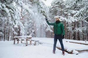 Portrait of smiling bearded male guy dressed in warm clothes, shakes tree covered with snow, has pleased expression, enjoys spending free time or weekends on fresh winter air. Season concept photo