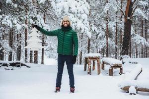 Full growth portrait of cheerful smiling male demonstrates white artificial fir tree, stands in winter forest has happy expression, glad to spend time outdoors. Winter and season concept photo