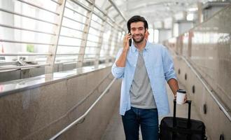 The man using the smartphone while the other hand holding a cup of coffee and pulling the luggage at the airport. photo