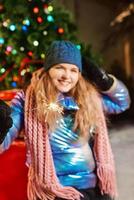 Happy smiling young Caucasian woman in scarf, hat, jacket, mittens with sparkler by the christmas tree outdoor. New year, fun, winter concept photo