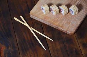 una toma detallada de un juego de rollos de sushi japoneses y un dispositivo para su uso de palillos, que se encuentran en una tabla de cortar de madera sobre una mesa en la cocina de un bar de sushi foto