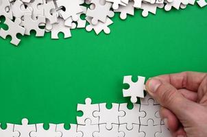 The hand folds a white jigsaw puzzle and a pile of uncombed puzzle pieces lies against the background of the green surface. Texture photo with space for text